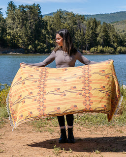 A woman standing outdoors by a lake holds up a large woven blanket or throw with a warm yellow base and rust-orange geometric floral patterns. Trees, water, and hills are visible in the background.