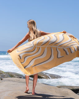 Woman holding a yellow and gray patterned towel on a rocky beach with ocean waves in the background