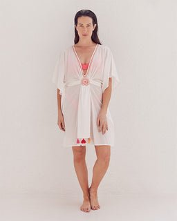 A female model wearing a white knee-length kaftan style cover up with a deep v-neck and multi-colored pick stitch details. The cover up has eye-catching front tassels and is tied at the waist.