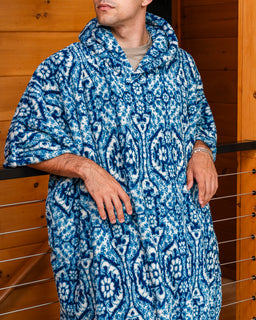 A man wearing a blue and white patterned hooded robe or poncho-style garment, standing indoors against a wooden wall with a railing visible behind him.