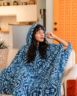 A woman wearing a blue and white patterned hooded blanket sits on a beige couch in a modern living room with a bright orange door in the background.