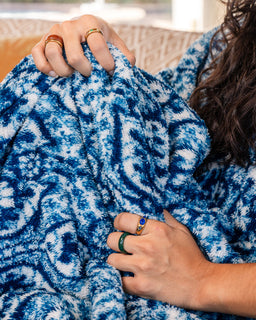A close-up of a person wrapped in a plush blue and white patterned blanket, wearing several rings on their hands.
