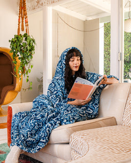 A woman sits on a beige sofa in a bright living room, wrapped in a plush hooded blanket that covers her entire body. She is holding an open book.