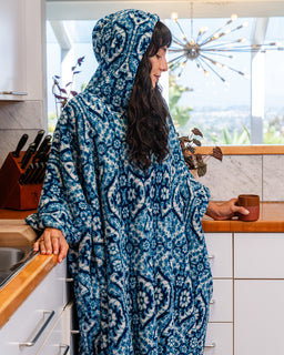 A woman wearing a blue and white patterned hooded robe stands in a modern kitchen, holding a brown ceramic mug.