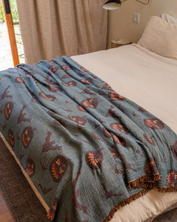 A blue-gray quilted blanket with a repeating jack-o'-lantern pattern in orange, brown, and purple, draped over a bed in a bedroom setting.