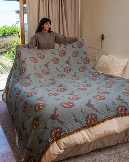 A woman holds up a large quilted blanket with a sage green background and a repeating pattern of brown silhouettes and orange circular motifs, shown on a bed in a bedroom setting.
