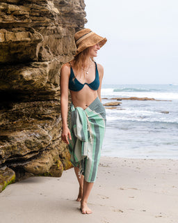 Woman in a bikini and sarong standing on a beach with ocean and rocks in the background