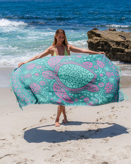A woman on a sandy beach holds up a large teal and pink patterned beach towel or wrap with a sea turtle design, with ocean waves and rocks in the background.