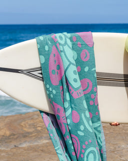 A lightweight patterned beach towel draped over a white surfboard on a rocky beach with the ocean in the background. The towel features a teal green base with pink and mint green paisley patterns.