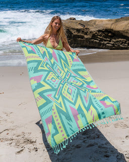 A woman on a sandy beach holds up a large patterned beach towel. The towel features a vibrant geometric design in shades of mint green, lime green, pink, and yellow, with matching mint green tassels along the edge.