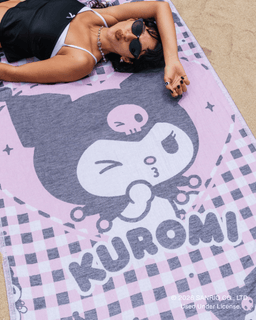 A woman lying on a pink and gray Kuromi beach towel on sand. The towel features a large Kuromi character graphic, checkered gingham borders, and the word 'KUROMI' printed across the bottom.