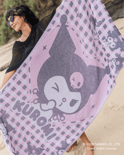 A woman on a beach holds up a large pink and lavender Kuromi-themed beach towel or blanket featuring Kuromi's face, checkered pattern, and the word 'KUROMI'.