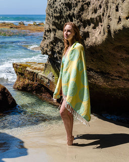Woman standing on a beach with a yellow and green patterned towel draped over her shoulders.