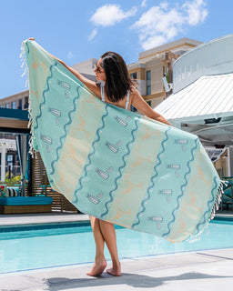 Woman holding a large turquoise towel by a pool with buildings in the background
