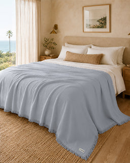 A light blue textured throw blanket with fringe edges draped over a bed in a warm, neutral-toned bedroom. The bed is styled with white sheets, a tan pillow, and a beige headboard, with a window showing a view of the ocean and palm tree in the background.