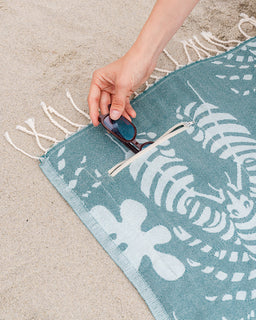 Person placing sunglasses into a teal beach towel with white patterns on sand