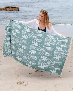 Woman holding a teal towel with white floral patterns on a beach.