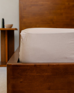 Wooden bed frame with a white fitted sheet in a minimalistic bedroom setting.