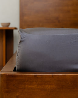 Gray pillow on a wooden bed with a wooden headboard and side table in the background.