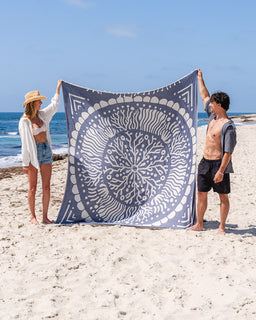 Two people holding a blue and white mandala-patterned towel on a beach.
