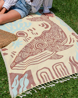 A woven outdoor towel with a stylized whale design in terracotta and light blue on a cream background, laid on green grass. A person wearing denim shorts and a striped shirt is partially visible sitting on the blanket, along with a woven bag