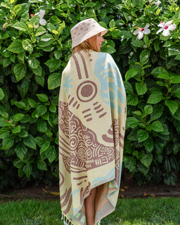 A woman stands outdoors in front of a leafy green hedge, wrapped in a cream-colored blanket with brown and light blue abstract patterns. She is wearing a white crochet bucket hat.
