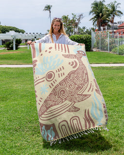 A woman outdoors on a grassy lawn holds up a large woven towel featuring a stylized whale design in brown, beige, and light blue. The background shows a sunny park with palm trees, a white fence, and a building.