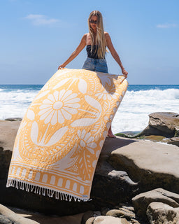 Woman holding a yellow and white patterned towel on a rocky beach with ocean waves in the background.
