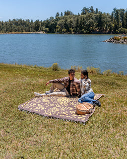 A couple sits on a patterned blanket on a grassy lakeside lawn, with a lake, trees, and blue sky in the background. A woven basket sits beside them.