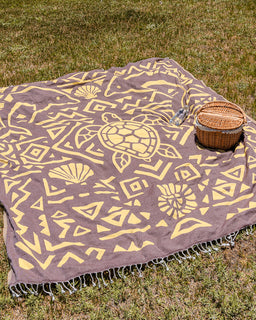 A large woven picnic blanket with a yellow tropical turtle and geometric pattern on a brown background, laid out on green grass next to a wicker basket and a clear water bottle.