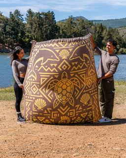 Two people standing outdoors by a lake holding up a large brown and tan patterned fabric item with a central turtle design and geometric motifs. Trees, water, and a dirt ground are visible in the background.