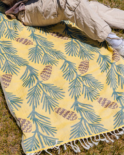 A yellow woven blanket with a pine branch and pinecone pattern, laid out on grass with a person's legs and shoes visible at the top edge.