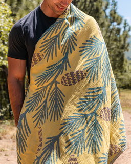 A man outdoors wearing a mustard yellow blanket or throw with a pine branch and pinecone pattern in blue-gray and brown.