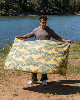 A man standing outdoors by a lake holds up a large blanket or sheet with a yellow background and a repeating pinecone and palm-leaf style print in muted blue-green and brown.