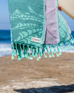 A close-up of a turquoise and lavender woven beach towel with tassels, held up outdoors with the ocean and sandy beach blurred in the background. A small woven label reading 'SAND CLOUD' is visible near the bottom.