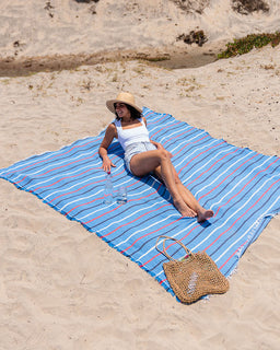Woman lying on a blue striped beach towel with a straw hat and bag on a sandy beach.