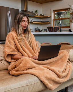 A woman sits on a beige sofa in a living room, wrapped in a plush tan blanket and using a black laptop. The background shows a kitchen area with wooden shelves, a stainless steel refrigerator, and a window.