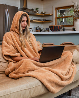 A woman sits on a beige sofa in a modern kitchen setting, wrapped in a plush, warm tan hooded blanket and using a black laptop.