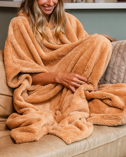 A woman sitting on a beige couch wrapped in a plush, warm tan throw blanket. The blanket has a soft, sherpa-like texture and is draped generously over her body.