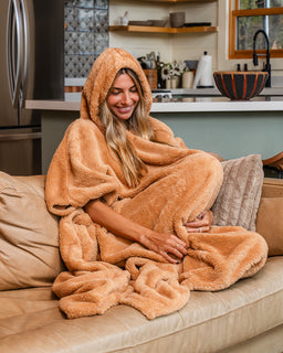 A woman sitting on a beige couch in a cozy living room, wrapped in a plush hooded blanket that resembles a teddy bear. The blanket is a warm camel brown color and has a soft, fuzzy texture.