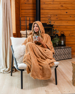 A woman sits in a cushioned armchair wrapped in a plush hooded blanket inside a rustic wood-paneled room, holding a mug.