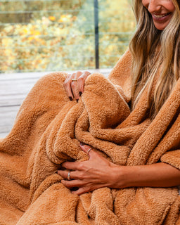 A woman wrapped in a plush, warm tan blanket on a wooden deck with a blurred outdoor background.