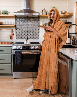 A woman standing in a kitchen wearing a large tan hooded blanket wrap and holding a mug.