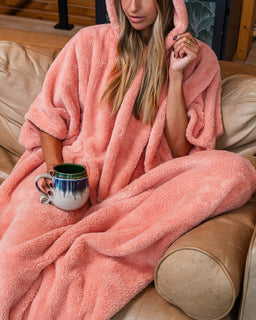 A woman sitting on a tan leather couch wrapped in a plush hooded blanket, holding a ceramic mug. The blanket is a soft dusty pink color.