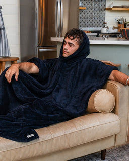 A man wearing a plush navy blue hooded blanket poncho sits on a beige leather couch in a modern living room setting.