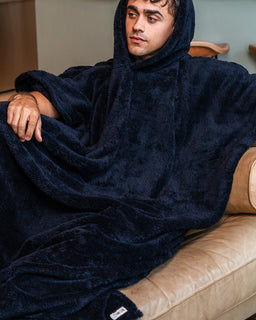 A man wearing a plush navy blue hooded blanket poncho sits on a beige leather couch in a living room setting.