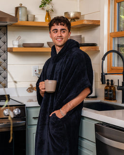 A smiling man wearing a plush navy blue hooded robe stands in a modern kitchen, holding a beige ceramic mug.
