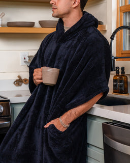 A man wearing a plush navy blue hooded robe stands in a kitchen, holding a beige ceramic mug. The robe has a soft, fuzzy texture and a large hood, with a pocket on the side.