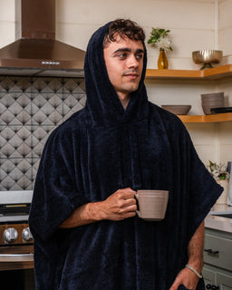 A man wearing a plush navy blue hooded bathrobe or poncho, holding a beige ceramic mug in a modern kitchen setting.