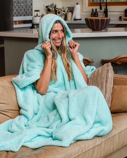 A woman sitting on a tan leather couch wrapped in a plush hooded blanket, smiling and holding the hood up. The blanket is a light aqua/mint color, and the background shows a modern kitchen and living room setting.